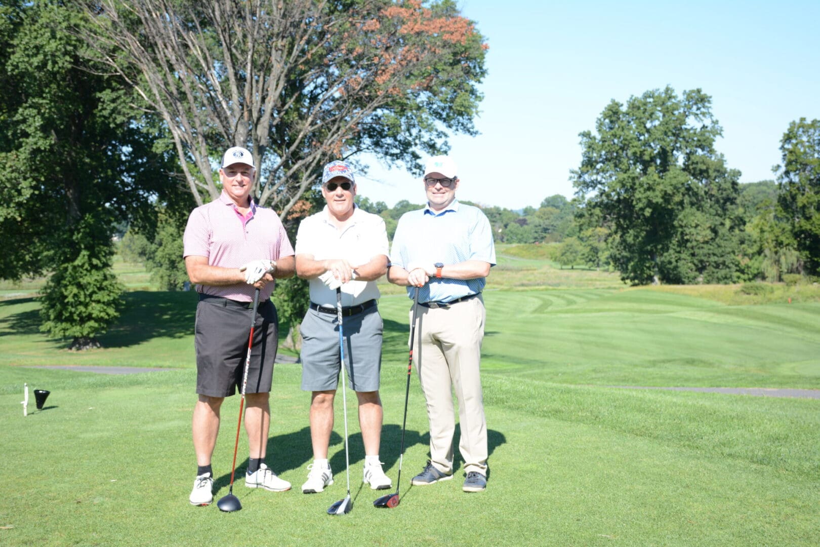 three men wearing polo shirt holding golf clubs in front of big tree
