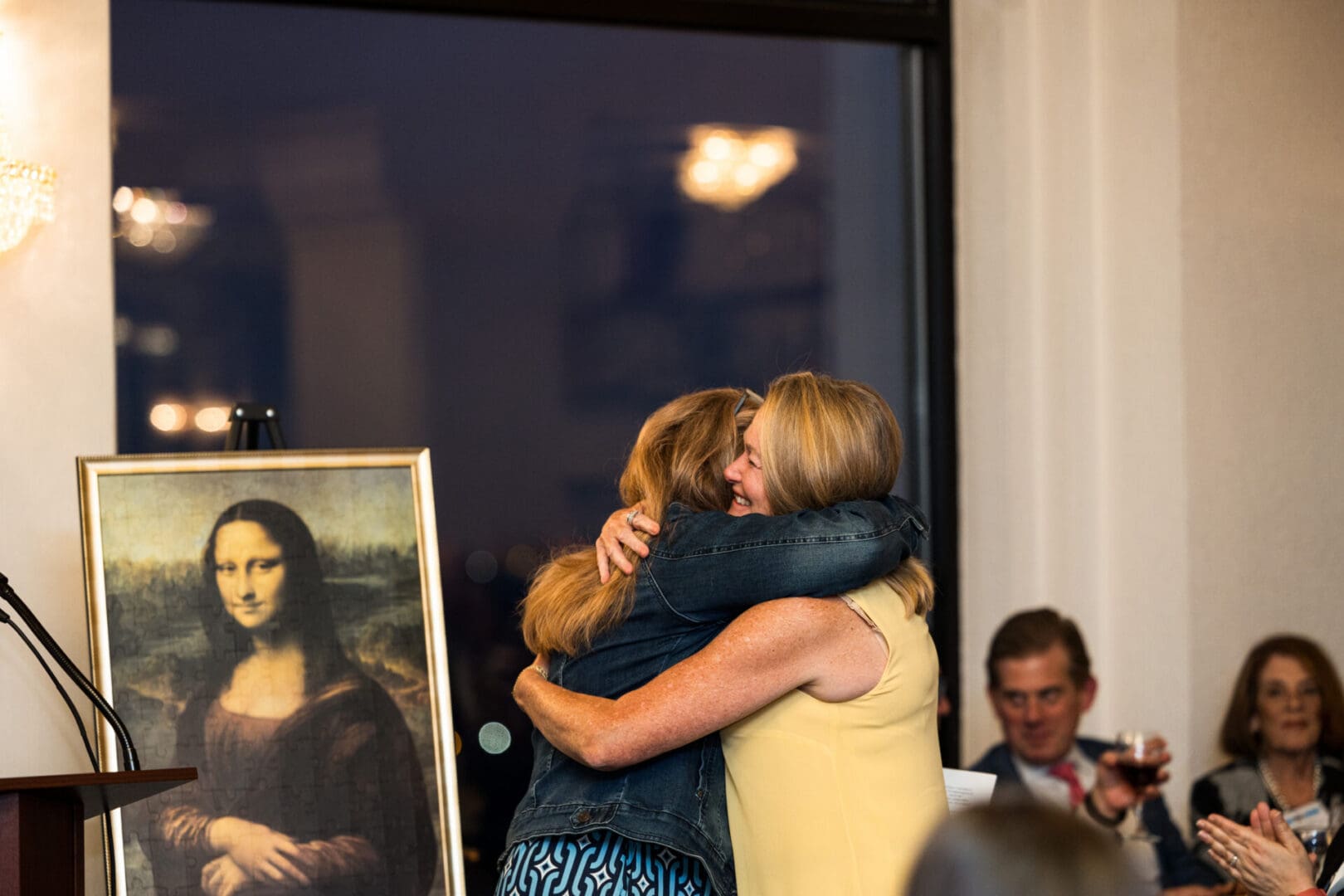 Two women hugging in front of a Mona Lisa painting.